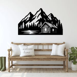 May include: Black metal wall art showing a mountain range, forest, and cabin scene. The artwork is mounted above a wooden bench with pillows that read "welcome home". Green wellington boots and a potted plant are also in the image.