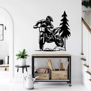 May include: Black and white silhouette of a motocross rider jumping over a hill with a pine tree in the background. The rider is wearing a helmet and goggles. The motorcycle has a large front tire and a smaller rear tire.