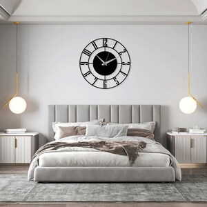 K&ouml;nnte beinhalten: Ein Schlafzimmer mit einem grauen, gepolsterten Bett, zwei wei&szlig;en Nachttischen und einer schwarzen Uhr mit r&ouml;mischen Ziffern an der Wand. Die Uhr hat ein schwarzes Zifferblatt und silberne Zeiger.
