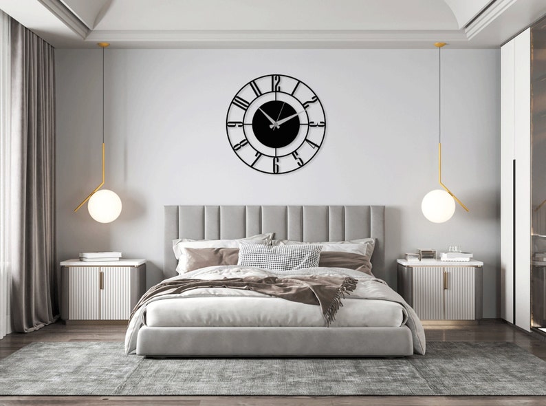 K&ouml;nnte beinhalten: Ein Schlafzimmer mit einem grauen, gepolsterten Bett, zwei wei&szlig;en Nachttischen und einer schwarzen Uhr mit r&ouml;mischen Ziffern an der Wand. Die Uhr hat ein schwarzes Zifferblatt und silberne Zeiger.