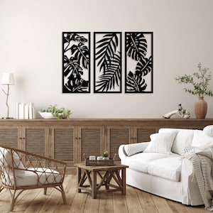 May include: Three black metal wall decor panels featuring a tropical leaf design. The panels are arranged horizontally on a white wall above a wooden cabinet and a white sofa.