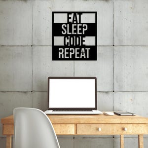 Puede incluir: Una pieza de arte mural de metal negro con las palabras "EAT SLEEP CODE REPEAT" en formato apilado. El arte cuelga sobre un escritorio de madera con una computadora portátil, un ratón y un cuaderno. Una silla blanca está frente al escritorio.