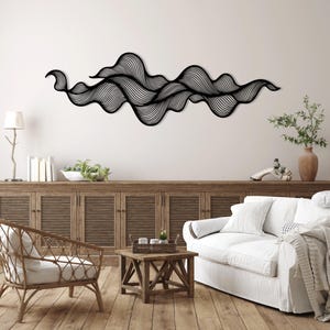 Op de afbeelding: Zwarte metalen wandkunst met een abstract ontwerp van gelaagde, gebogen lijnen. Het kunstwerk is bevestigd aan een witte muur boven een houten kast, wat een moderne uitstraling aan de kamer geeft.