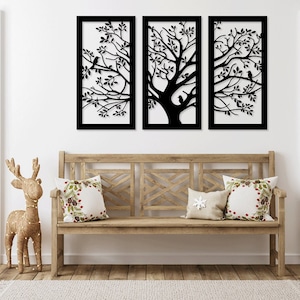 May include: Three black metal wall art pieces depicting a tree with birds. The tree is in silhouette and the pieces are arranged horizontally. The art is hanging on a white wall above a wooden bench with cushions.