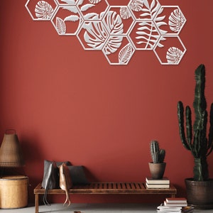 May include: A set of white hexagonal wall decor with a leafy design. The decor is arranged in a staggered pattern on a red wall. The decor is in a living room setting with a wooden bench, a cactus, and a rug.