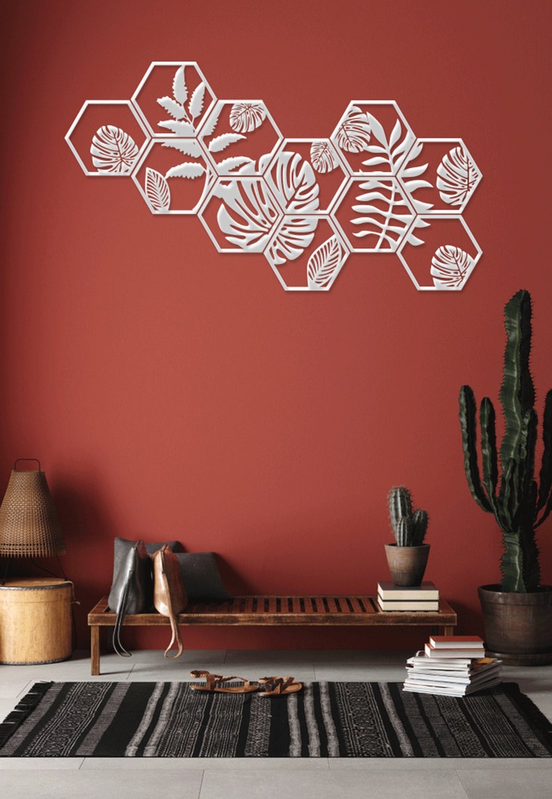 May include: A set of white hexagonal wall decor with a leafy design. The decor is arranged in a staggered pattern on a red wall. The decor is in a living room setting with a wooden bench, a cactus, and a rug.