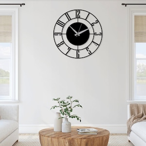 K&ouml;nnte beinhalten: Eine schwarze Wanduhr aus Metall mit r&ouml;mischen Ziffern und einem schwarzen Zifferblatt. Die Uhr h&auml;ngt an einer wei&szlig;en Wand in einem Wohnzimmer.