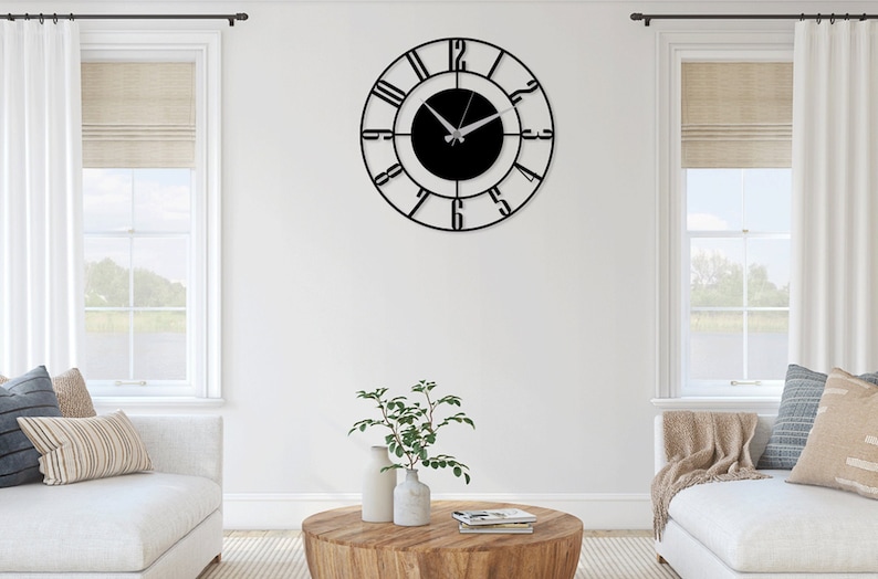 K&ouml;nnte beinhalten: Eine schwarze Wanduhr aus Metall mit r&ouml;mischen Ziffern und einem schwarzen Zifferblatt. Die Uhr h&auml;ngt an einer wei&szlig;en Wand in einem Wohnzimmer.
