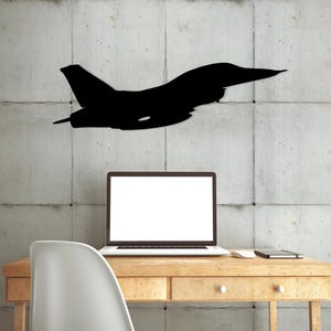 May include: A black silhouette of a fighter jet on a gray concrete wall, with a laptop computer and a white chair in front of a wooden desk.