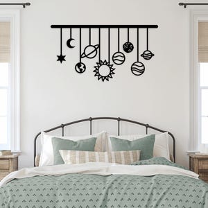 May include: Black metal wall art featuring celestial bodies, including a star, moon, sun, planets, and Earth. The decorative piece hangs above a bed with a green and white patterned comforter, creating a whimsical bedroom theme.
