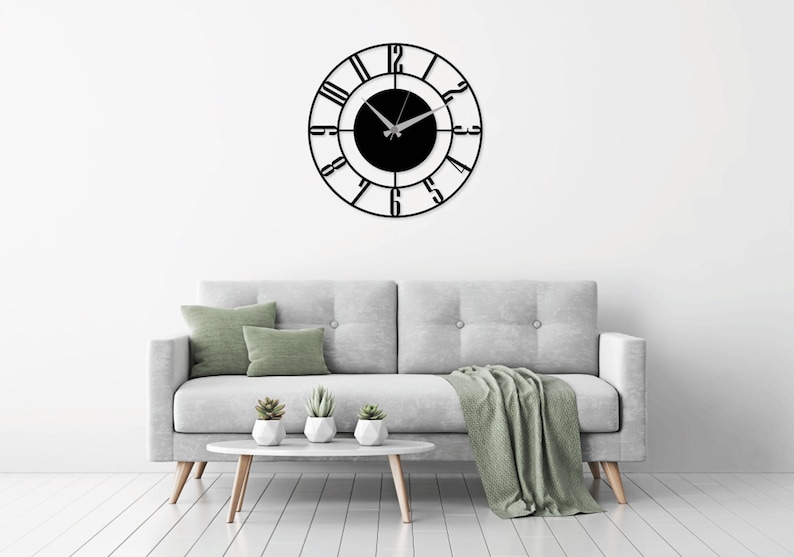 K&ouml;nnte beinhalten: Eine schwarze Metalluhr mit r&ouml;mischen Ziffern auf wei&szlig;em Hintergrund. Die Uhr h&auml;ngt an einer wei&szlig;en Wand &uuml;ber einem grauen Sofa mit gr&uuml;nen Kissen und einer gr&uuml;nen Decke.