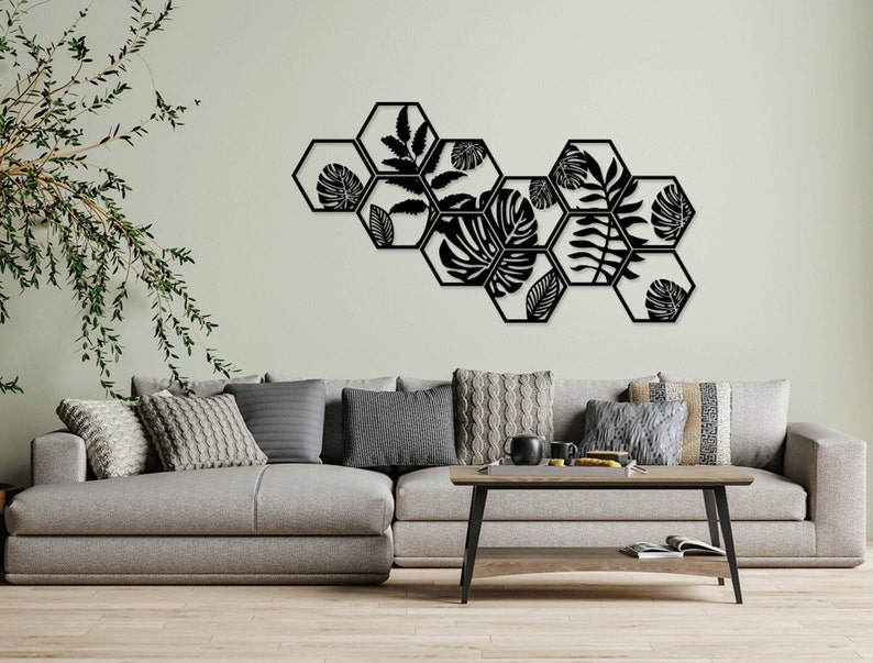 May include: Black metal wall decor featuring a geometric design of hexagonal shapes with cut-out leaf patterns. The decor is mounted on a light green wall above a grey sofa with cushions.