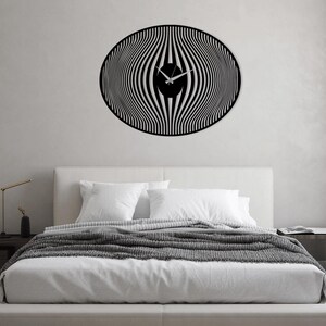 May include: A black and white striped wall clock with a modern design. The clock is hanging on a white wall above a white bed with a gray blanket.