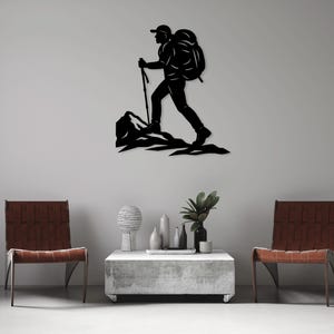 Puede incluir: Arte de pared de metal negro que representa a un excursionista con una mochila y un bastón de senderismo, ascendiendo una montaña. La silueta está sobre una pared neutra, con dos sillas y una mesa de centro moderna.