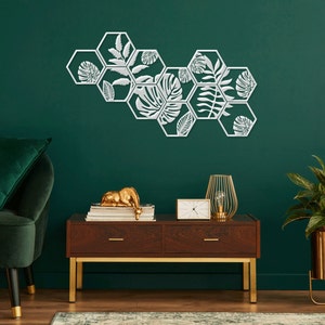 May include: A set of six white metal hexagonal wall decor pieces with a leafy design. The decor is mounted on a dark green wall. A green velvet sofa, a wooden table with gold legs, a gold plant stand with a green plant, and a gold clock are also in the image.