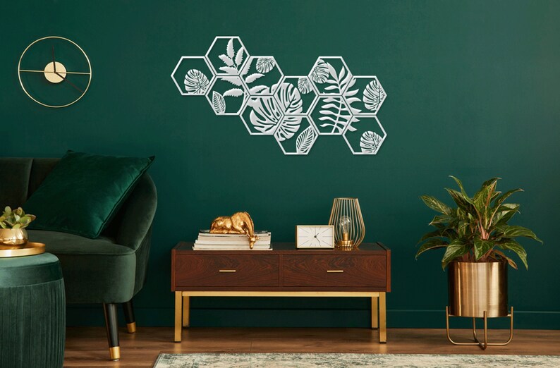 May include: A set of six white metal hexagonal wall decor pieces with a leafy design. The decor is mounted on a dark green wall. A green velvet sofa, a wooden table with gold legs, a gold plant stand with a green plant, and a gold clock are also in the image.