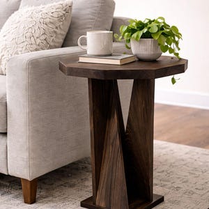 May include: Dark brown wooden side table with a geometric base. The tabletop holds a white mug, a book, and a small potted plant. The table is next to a gray sofa on a patterned rug.