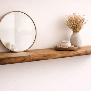 May include: A wooden floating shelf with a natural edge, holding a round gold-framed mirror, a vase of dried flowers, a small ceramic bowl, and a wooden tray with beads. The shelf is a warm brown color.