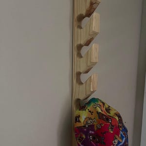 May include: A vertical light-colored wooden wall rack featuring four angled pegs for hanging items. A colorful baseball cap with a repeating cartoon character print in shades of red, yellow, purple, and blue hangs from the bottom-most wooden peg.