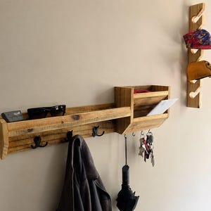 May include: A rustic wooden wall-mounted organizer with a shelf, hooks, and a mail slot. Sunglasses, a phone, and keys are visible. A separate wooden rack displays two baseball caps. The organizer is made of light-colored wood.