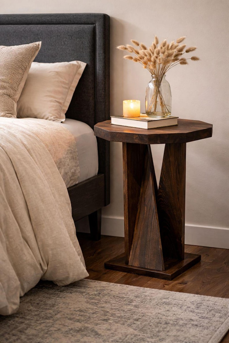 Small wooden side table styled as handmade bedside table, dark geometric base beside bed with dried bouquet.