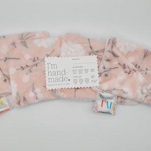 Puede incluir: Se muestran cuatro artículos de felpa rosa con estampado floral. Los artículos tienen un diseño floral blanco con tallos y hojas grises. Una etiqueta blanca dice "I'm handmade. Handle with care." Se adjuntan pequeñas etiquetas con las letras "IM".