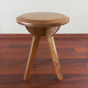 Puede incluir: Un taburete de madera con un asiento redondo y tres patas. El taburete está hecho de madera marrón claro y tiene un acabado natural.