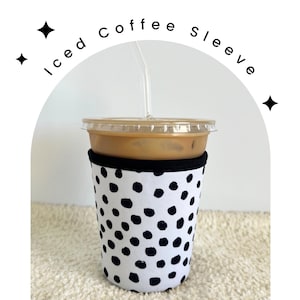 May include: A black and white polka dot coffee sleeve for an iced coffee cup. The sleeve is made of neoprene and has a black band around the top.