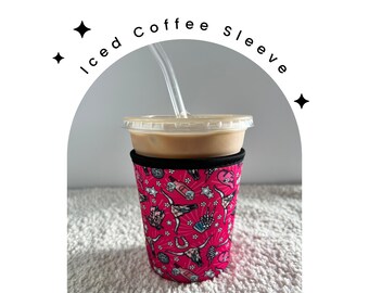 May include: Pink and black insulated coffee sleeve with a colorful pattern of cowboy boots, hats, and horses. The sleeve is on a white iced coffee cup with a straw.