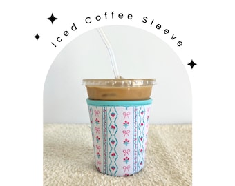 May include: A white and blue patterned coffee sleeve with pink flowers and bows. The sleeve is on a white iced coffee cup with a straw.