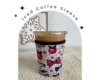 May include: A white and pink coffee sleeve with a design of red apples, stars, and a black bow. The sleeve is on a clear plastic cup with a brown iced coffee drink inside.