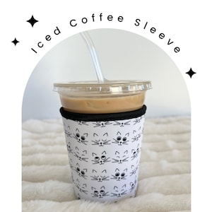May include: A white coffee sleeve with a black and white cat face pattern. The sleeve is on a white fuzzy surface and has a clear plastic cup with iced coffee inside.