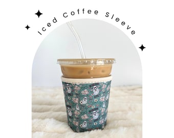 May include: A green and white coffee sleeve with a pattern of ghosts, glasses, and books. The sleeve is on a white fuzzy surface and has a clear plastic cup with iced coffee inside.