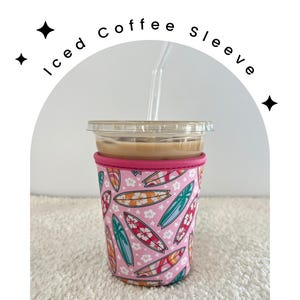May include: Pink and white coffee sleeve with a surfboard pattern. The sleeve is made of neoprene and has a pink trim. The sleeve is on a white surface.