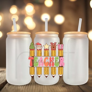 May include: Three frosted glass tumblers with bamboo lids and silver straws. The center tumbler features a colorful design with Christmas-themed icons atop pencils and the word "TEACHER". The design includes a Christmas tree, Santa, a star, a reindeer, and a gift.