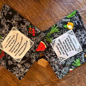May include: Two square fabric pot holders with a black background featuring white floral line art and colorful fruit illustrations. Each features a white center patch with printed Bible verses: Matthew 7:7 and 2 Kings 20:5, with black stitched borders.