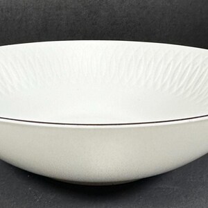 May include: A white ceramic serving bowl with a black rim. The interior features a subtle, textured wave pattern. The bowl is round and has a wide, shallow design, suitable for salads or other dishes.