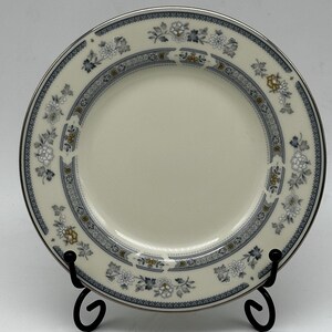 May include: A white ceramic plate with a blue and gold floral pattern around the rim. The plate is on a black stand.