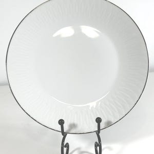 May include: A white ceramic plate with a silver rim and a subtle, repeating leaf pattern. The plate is displayed on a black metal stand, showcasing its design. This elegant plate is suitable for serving food.