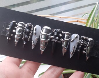 Puede incluir: Un conjunto de diez uñas postizas negras y plateadas con varios diseños, incluyendo pinchos, tachuelas y cadenas.