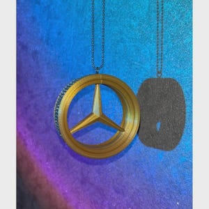 May include: A gold-colored pendant featuring a three-pointed star emblem within a circular frame. The pendant hangs from a silver-colored chain. The background is a gradient of blue, purple, and black.