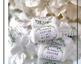 May include: White wrapped candies with labels that read "The Wedding Of OLIVER and KERRY 10/09/26". The labels have a floral design. The candies are on a reflective surface with white flowers in the background.