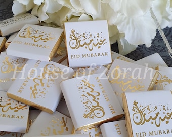 Puede incluir: Un montón de barras de chocolate blanco con envoltorios de papel de aluminio dorado. Los envoltorios tienen caligrafía árabe en dorado que dice "Eid Mubarak".