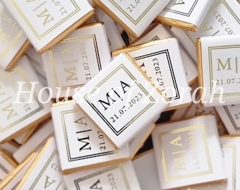 May include: A close-up of a pile of small, white chocolate bars with gold foil wrappers. Each bar has a black border and the name "MIA" printed in black with the date "21.07.2023" printed below.