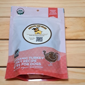 May include: A red and white resealable bag of organic turkey jerky dog treats. The bag features a cartoon turkey and the text "Organic Turkey Jerky Recipe Bites for Dogs made with organic sweet potatoes." The bag also has a USDA Organic seal and a UPC code.
