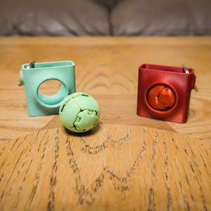 May include: Two square, small containers, one mint green and one red, with a circular opening and a metal clasp. A green, segmented ball sits between them. The objects are on a wooden surface.