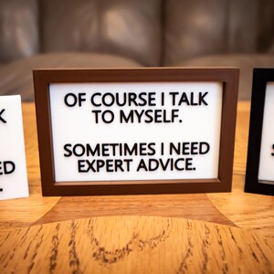 May include: Three framed signs with the text "OF COURSE I TALK TO MYSELF. SOMETIMES I NEED EXPERT ADVICE." and similar phrases. The frames are brown, black, and white, set on a wooden surface.