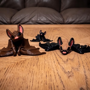 May include: Three 3D printed bat figurines. One is brown with pink ears, one is brown with black wings, and one is black with brown wings.