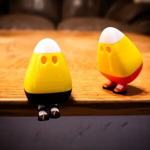 May include: Two small, three-dimensional figures shaped like candy corn. Each figure has a white top, a yellow middle section, and a base with small brown shoes. The figures feature two circular indentations for eyes on the front yellow surface.