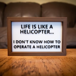 May include: A framed sign with the text "LIFE IS LIKE A HELICOPTER... I DON'T KNOW HOW TO OPERATE A HELICOPTER" in black letters. The frame is brown, and the background is a blurred brown leather couch.